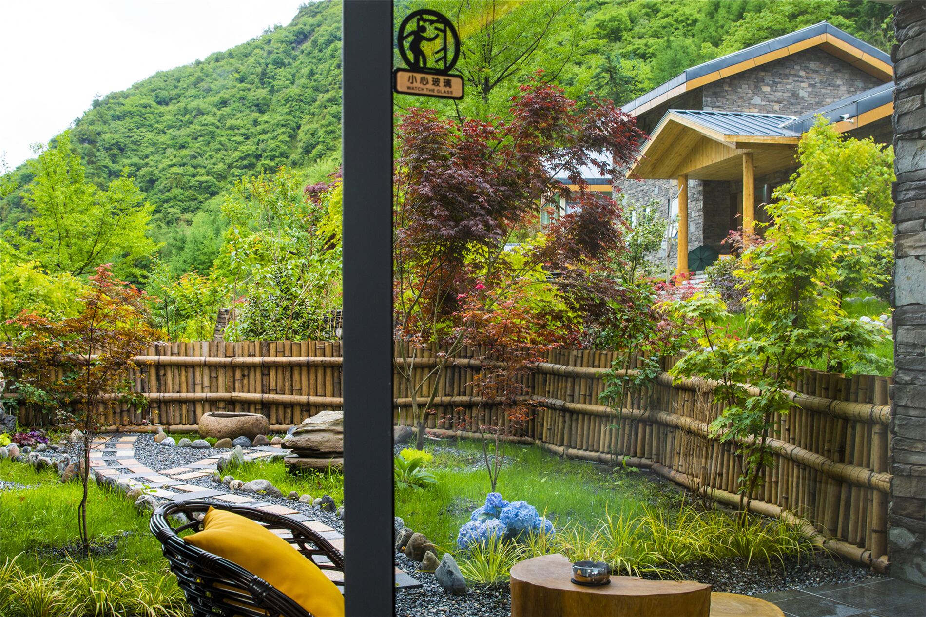 A garden with a bamboo fence, a stone building, and a sign with a person and a glass.