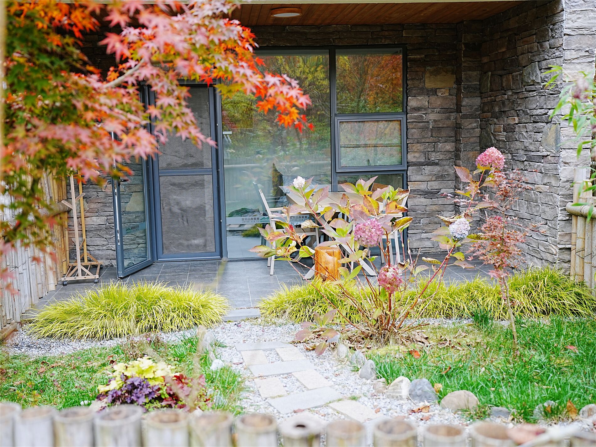 A stone building with a glass door, a wooden deck, and a garden with various plants and a pathway.