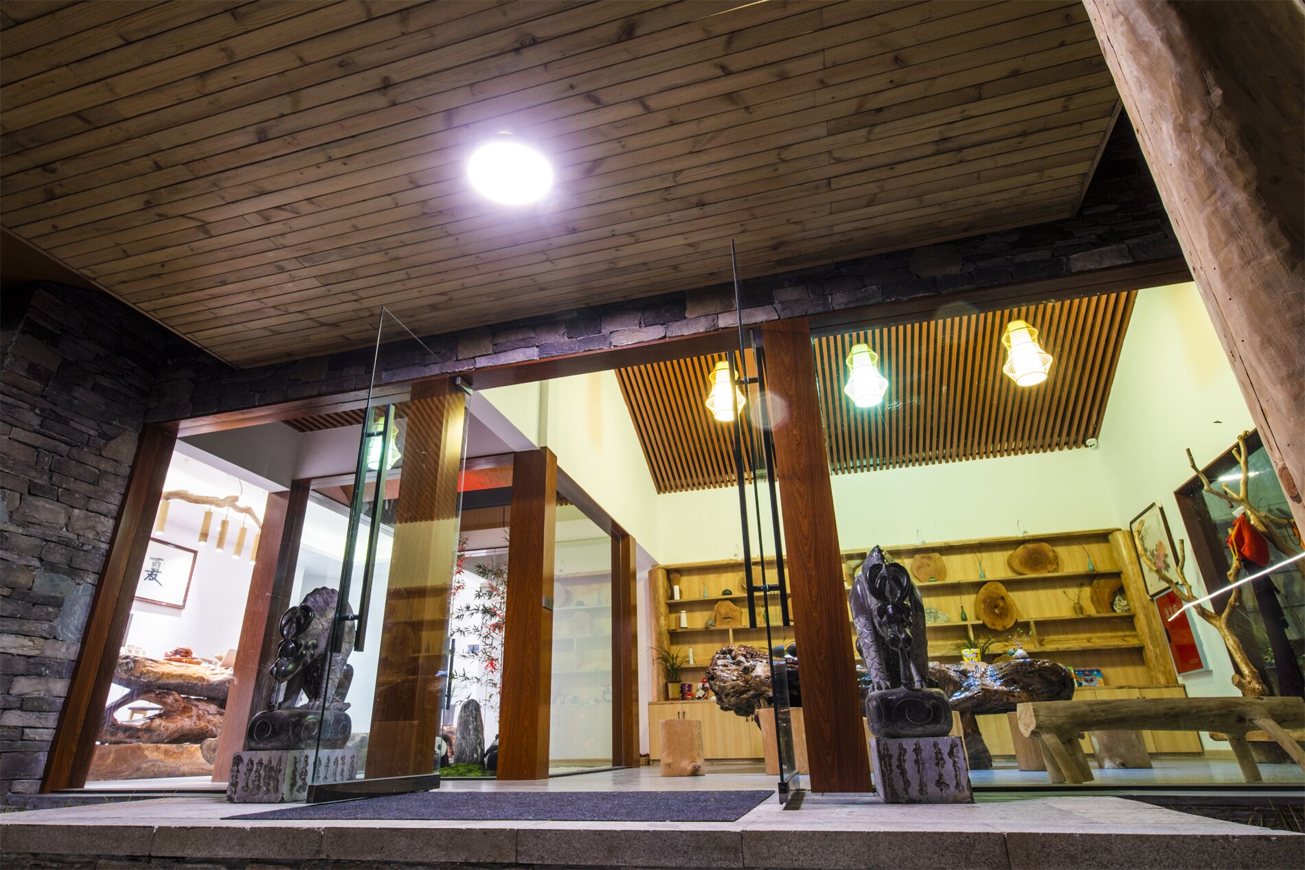 A room with wooden ceiling beams, stone walls, and a glass partition. There are wooden sculptures on pedestals and a wooden bench.