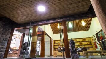 A room with wooden ceiling beams, stone walls, and a glass partition. There are wooden sculptures on pedestals and a wooden bench.