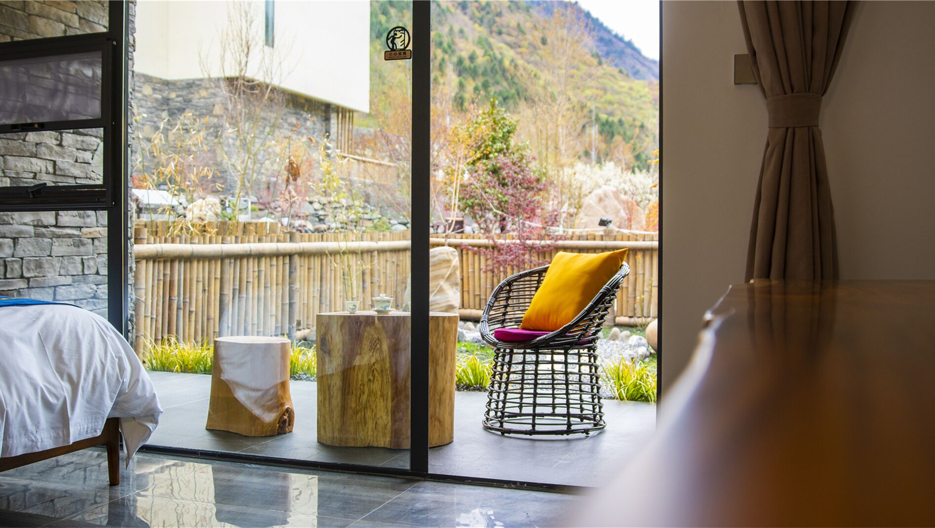 A room with a bed, a chair with a cushion, a small table, and a view of a garden and mountains.