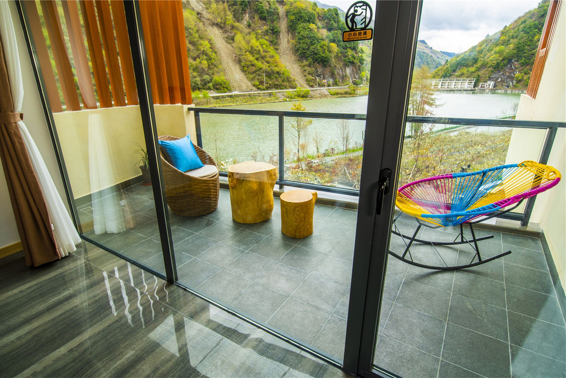 Un balcón con mobiliario de mimbre, una silla colgante colorida y vistas a un río y colinas.