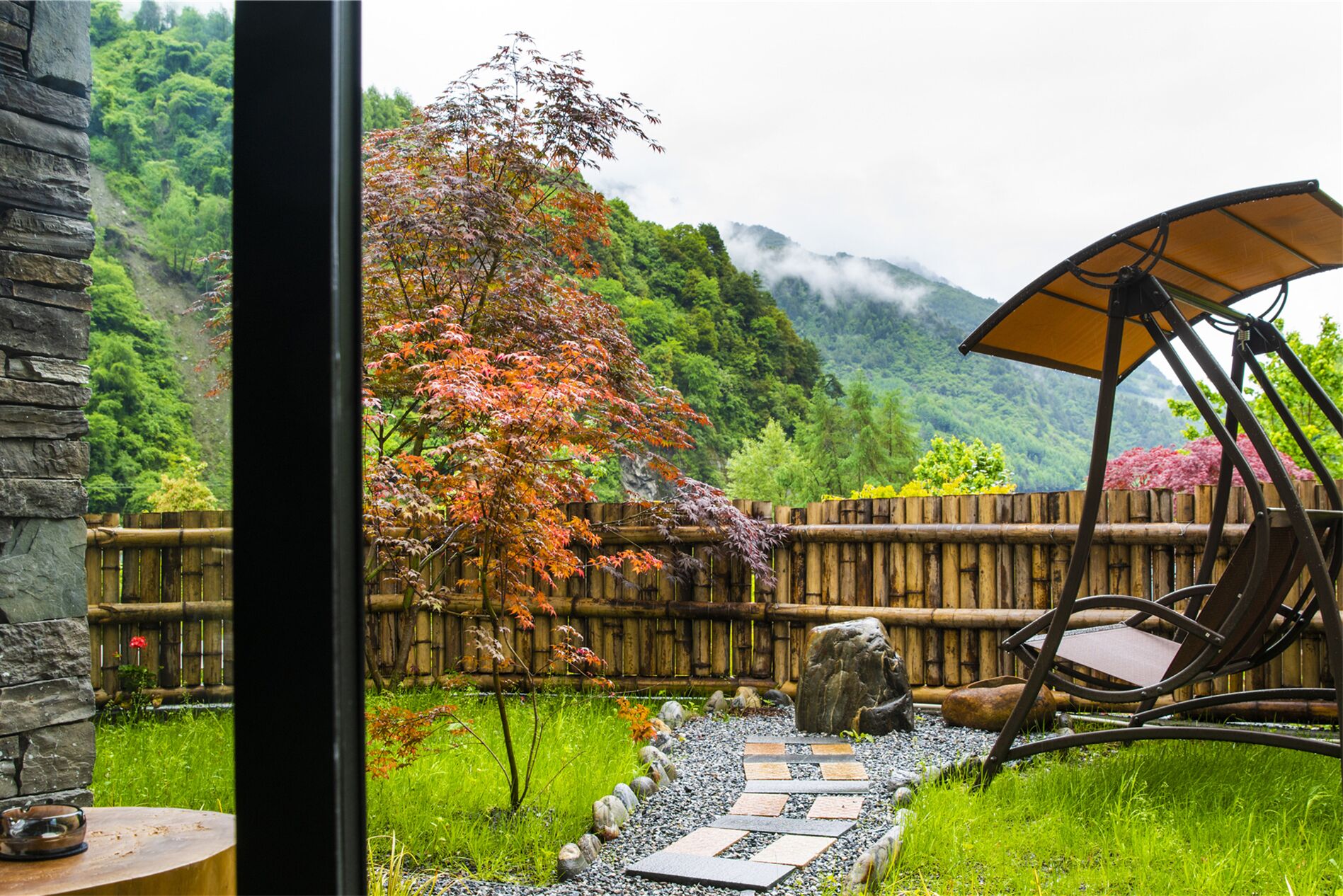 A view through a window of a garden with a bamboo fence, a stone pathway, and a chair with a canopy.