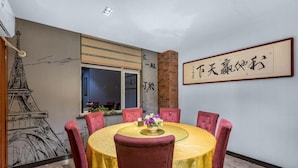 Un coin repas avec une table ronde, des chaises à revêtement rouge et une nappe jaune. Un mur est décoré d’une fresque représentant la Tour Eiffel, et au-dessus de la table, on peut voir une œuvre de calligraphie chinoise encadrée.