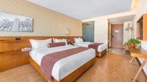Une chambre d’hôtel avec deux lits, une tête de lit en bois, une table de chevet, une petite table, une lampe fixée au mur, une grande fenêtre et une porte donnant sur une autre pièce.