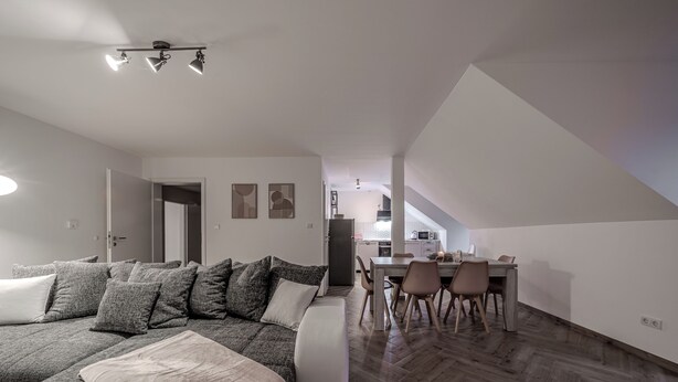 Un salon moderne avec un grand canapé d’angle, un coin repas avec une table et des chaises en bois, et une cuisine en arrière-plan.
