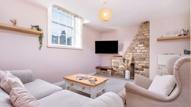 A living room with a sofa, armchair, and a coffee table. There is a TV mounted on the wall, a stone fireplace, and a window with a view of buildings outside. - Stone cottage - In the Heart of Stamford (Stamford)