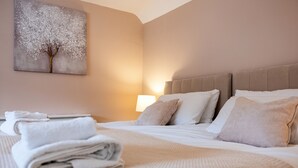 A neatly made bed with white linens and a folded towel, a beige headboard, and a wall-mounted painting of a tree. - Stone cottage - In the Heart of Stamford (Stamford)