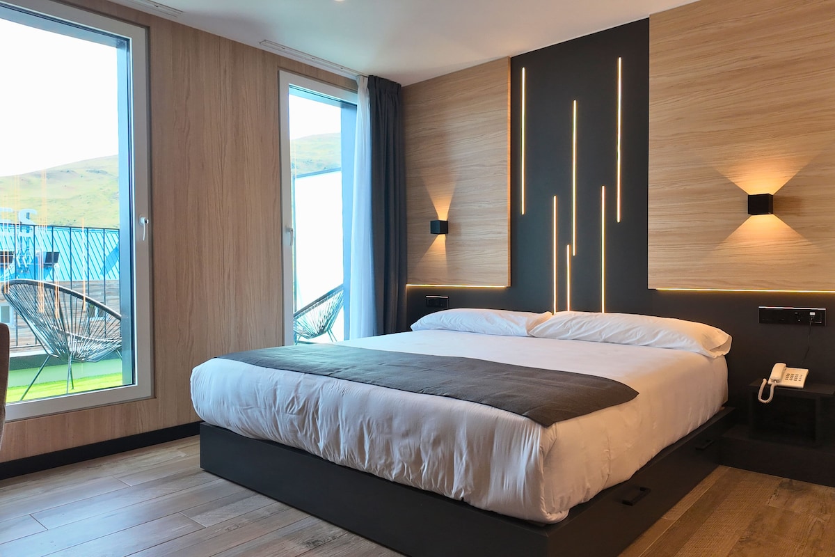 Una camera d'albergo moderna con un letto grande, vista sull'esterno, una sedia e un telefono sul comodino.