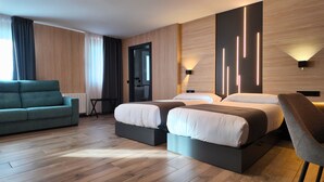 A modern hotel room with a sofa, two beds, a small table, and a bathroom visible through an open door.