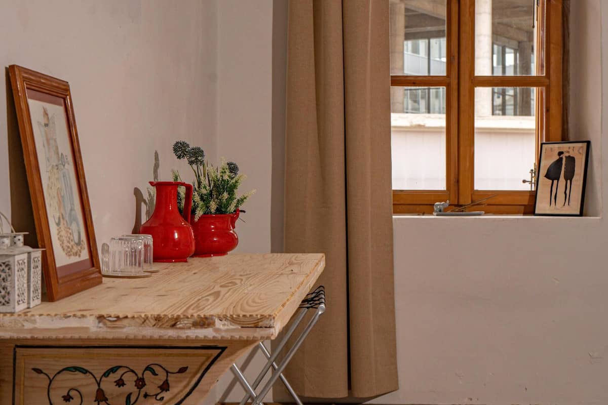 A room with a wooden desk, a mirror, a red vase with flowers, a framed picture, and a window with curtains.