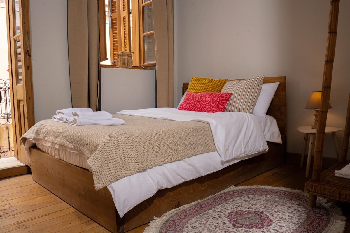 A wooden bed with white bedding and multiple pillows, a small round rug, and a wooden nightstand with a lamp.
