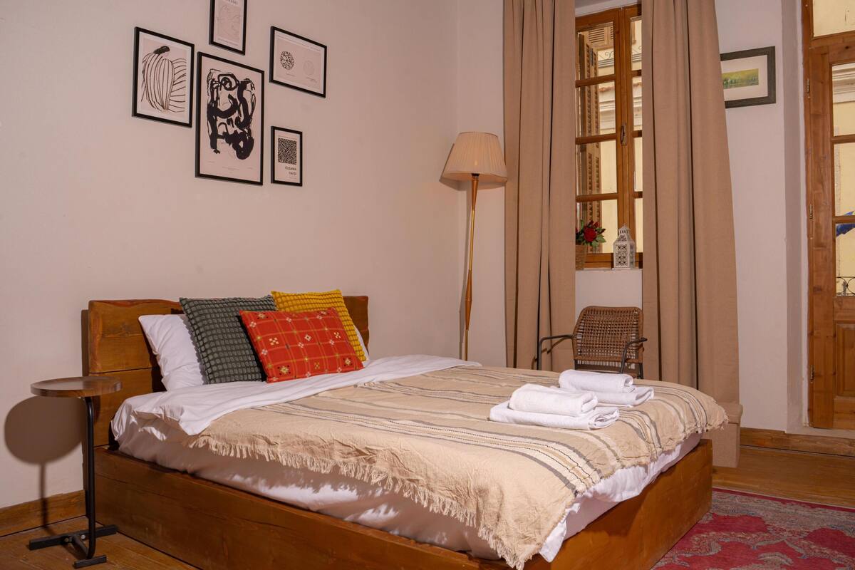 A neatly made bed with a striped blanket, a wooden headboard, and multiple pillows. There is a chair and a lamp beside the bed.