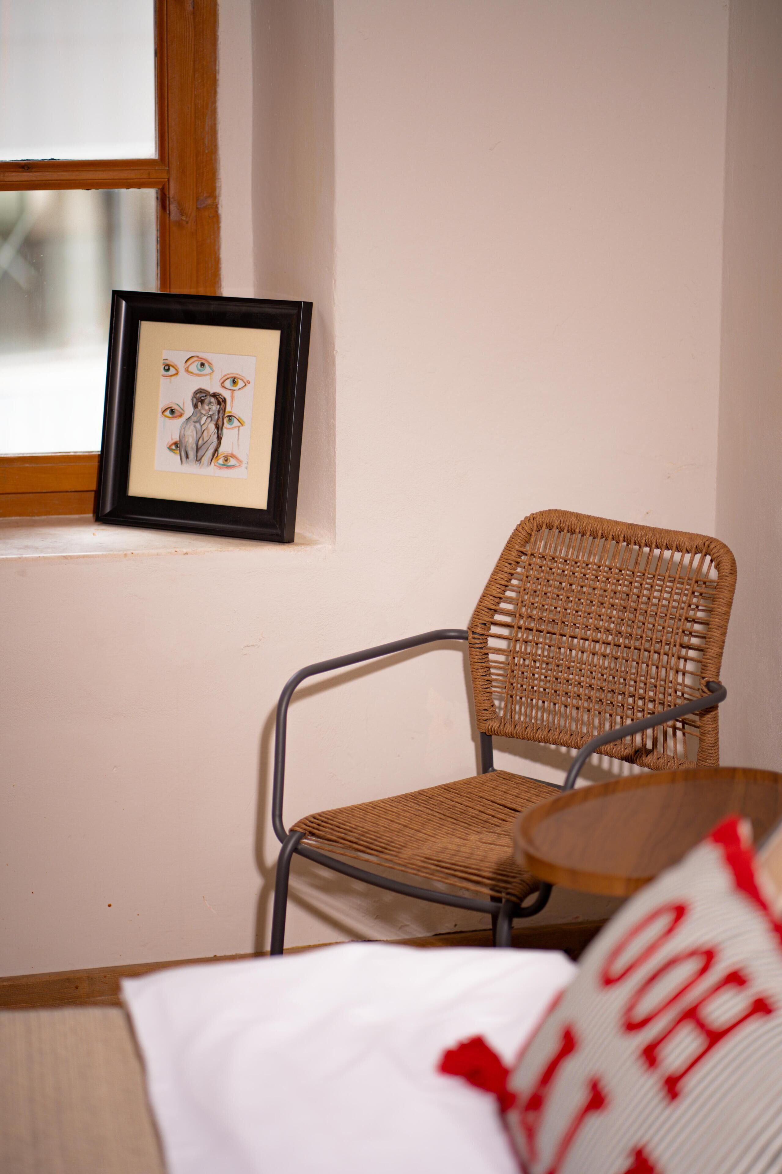 A wicker chair with a metal frame, a framed artwork on the wall, and a white pillow with red text.