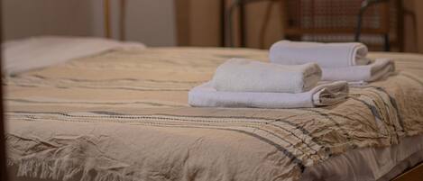 Ein ordentlich bezogenes Bett mit einer beige farbenen Decke und zwei gefalteten Handtüchern, eine Stehlampe und ein Sessel in einem Zimmer mit Fenster und einer Vase mit Blumen.