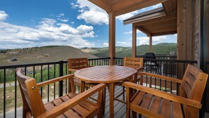 Une terrasse en bois avec une table ronde et des chaises, offrant une vue panoramique sur un paysage pittoresque composé de collines et d’un ciel dégagé.