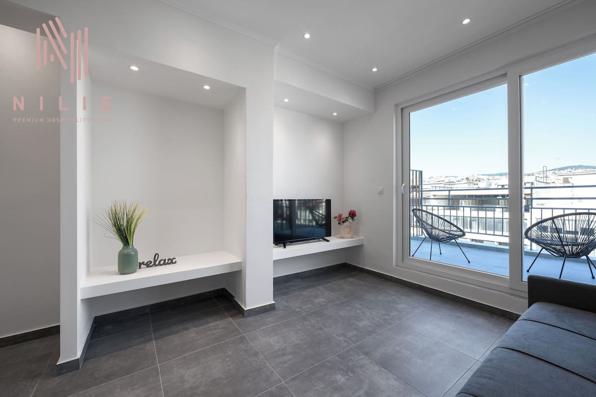 Un salon moderne avec un téléviseur à écran plat, un banc, une plante et deux fauteuils placés près d’une grande fenêtre offrant une vue sur la ville.