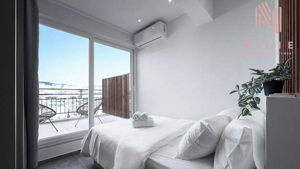 A modern hotel room with a large window, a bed with white linens, a potted plant, and a view of the city.