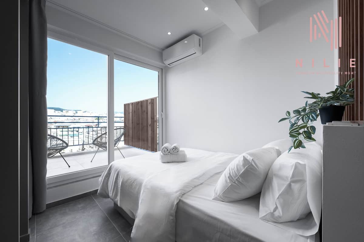 Une chambre d’hôtel moderne avec une grande fenêtre, un lit avec du linge de maison blanc, une plante en pot et une vue sur la ville.