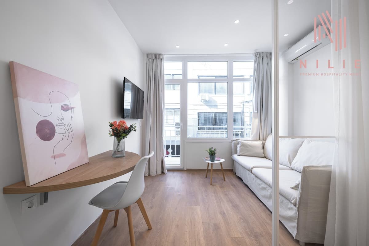 Une chambre d’hôtel moderne avec un bureau en bois, un canapé blanc, une petite table avec une plante et une grande fenêtre avec des rideaux.