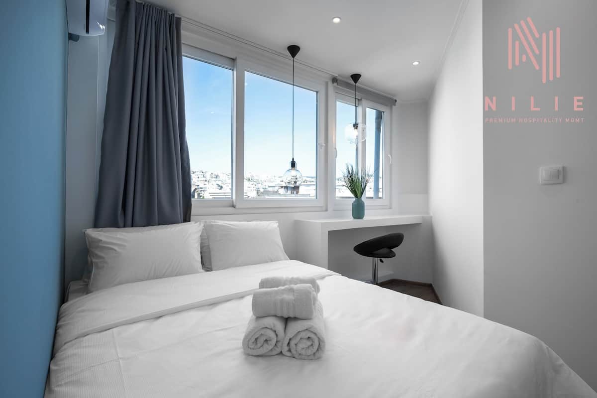 Une chambre d’hôtel moderne, dotée d’un grand lit, d’un bureau et offrant une vue sur la ville grâce à de larges fenêtres.