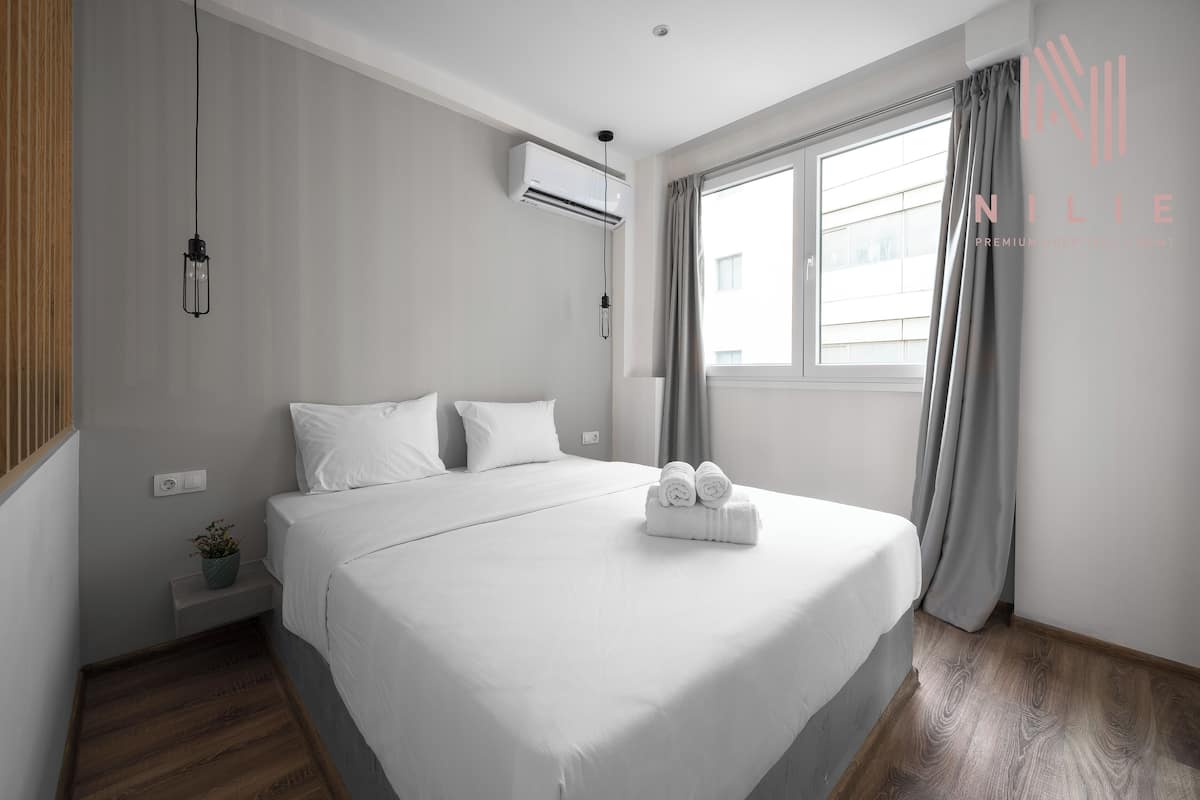 Une chambre d’hôtel moderne avec un grand lit, une petite table de chevet agrémentée d’une plante et une fenêtre avec des rideaux.