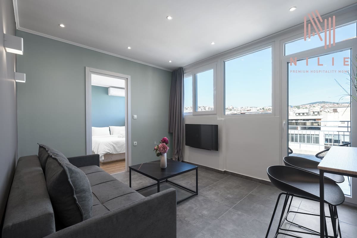 Une chambre d’hôtel moderne avec un canapé gris, une petite table ornée d’un vase de fleurs, un coin repas avec une table et des chaises, et une grande fenêtre donnant sur la ville.