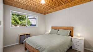 A bedroom with a wooden ceiling, a bed with a wooden headboard, a nightstand with a lamp, and a window with a view of greenery. - Pelican Point- Located next to Edwards Point Wildlife Reserve (St Leonards)