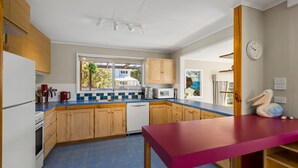 A compact kitchen with wooden cabinets, a red table, and a view of a house through the window. - Pelican Point- Located next to Edwards Point Wildlife Reserve (St Leonards)