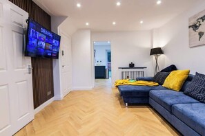 A modern living room with a television displaying a drama channel, a blue sofa with yellow and black cushions, a lamp, and a herringbone wooden floor. - Charming Home in Bromsgrove: Perfect for Groups (Birmingham)
