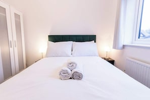 A neatly made bed with rolled towels, a green upholstered headboard, and bedside lamps. - Charming Home in Bromsgrove: Perfect for Groups (Birmingham)