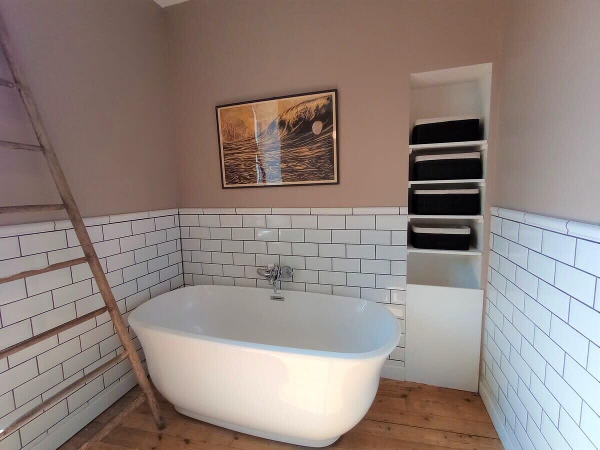 A bathroom with a freestanding bathtub, white tiled walls, a wooden floor, and a framed picture on the wall.