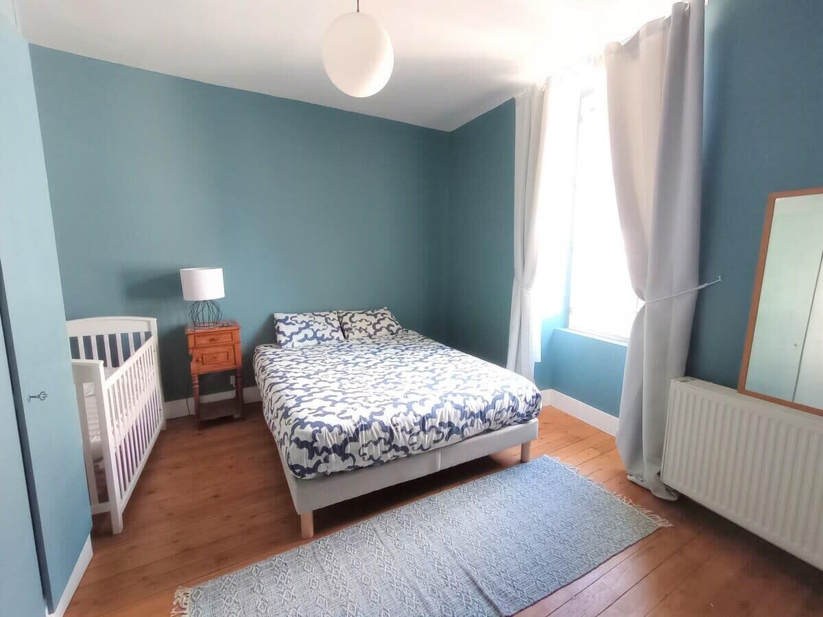 A bedroom with a bed, a nightstand, a lamp, a crib, and a window with curtains.