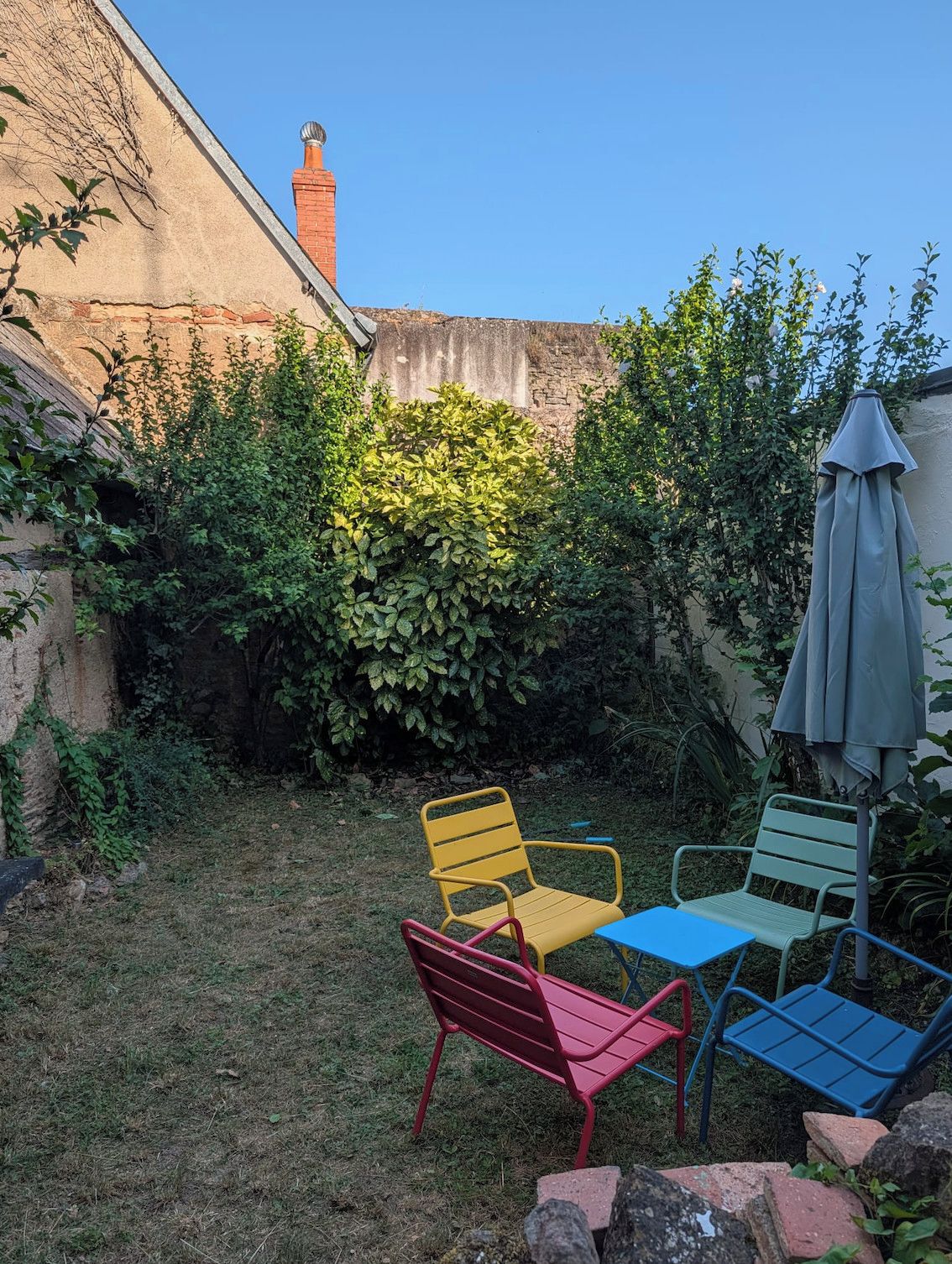 Ein Gartenbereich mit Backsteinmauer, Schornstein und verschiedenen Pflanzen. Bunte Metallstühle und ein Tisch sind ebenfalls vorhanden.