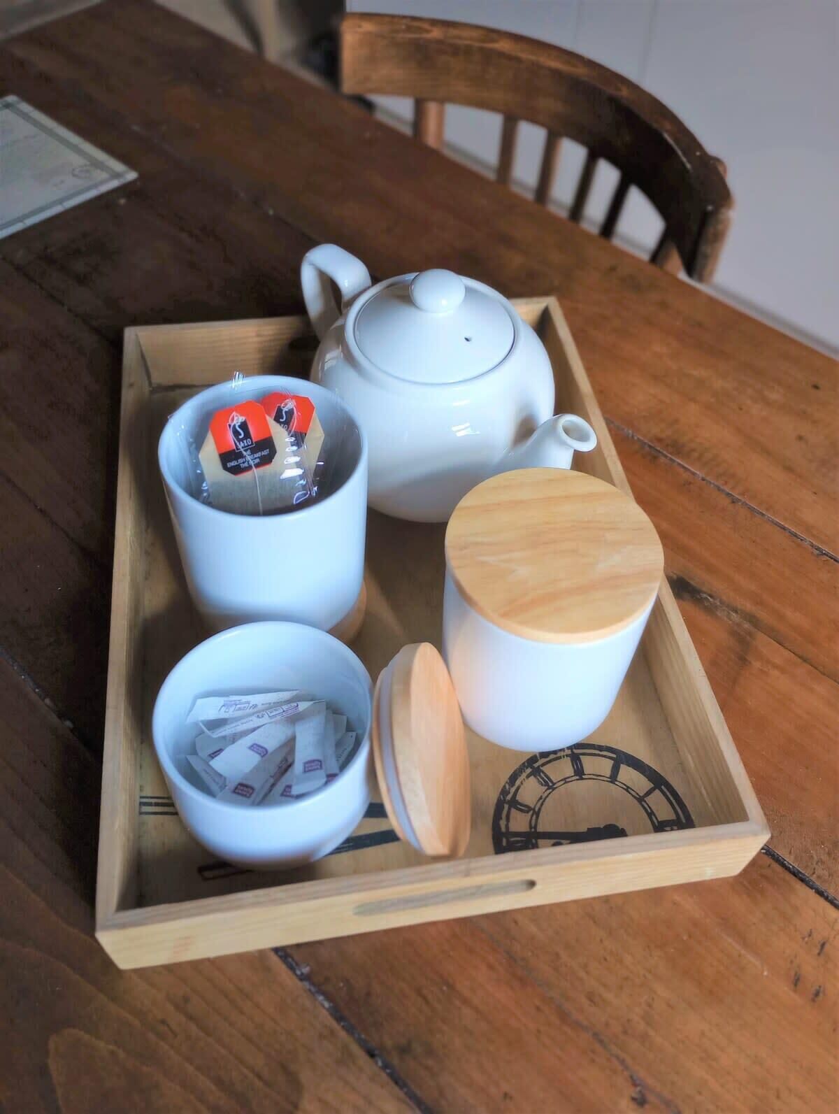 Ein Holztablett mit einer weißen Teekanne, einer weißen Tasse, einem Holzboden und einer kleinen Schale mit Teebeuteln.