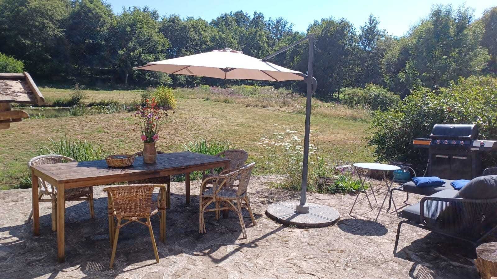Une terrasse extérieure avec une table en bois et des chaises en osier, un grand parasol et un barbecue.