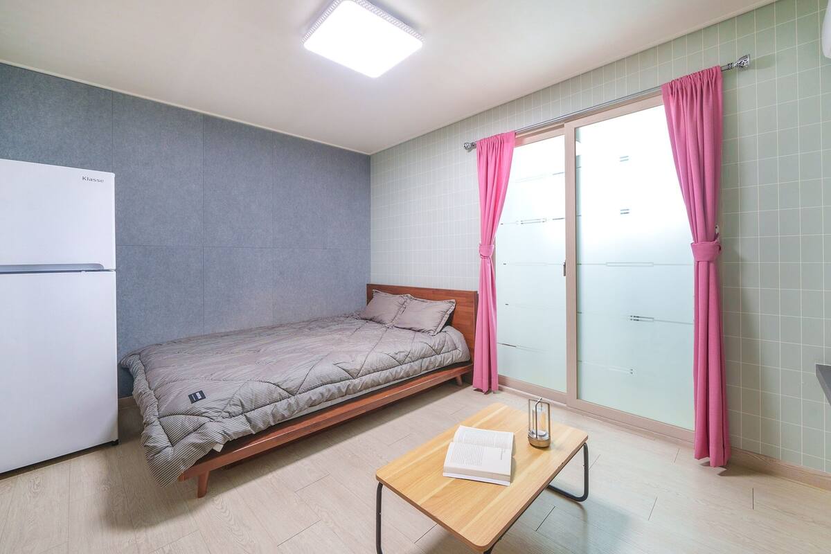 Een kleine slaapkamer met een bed, een tafeltje, een witte koelkast, een raam met gordijnen en een potplant.