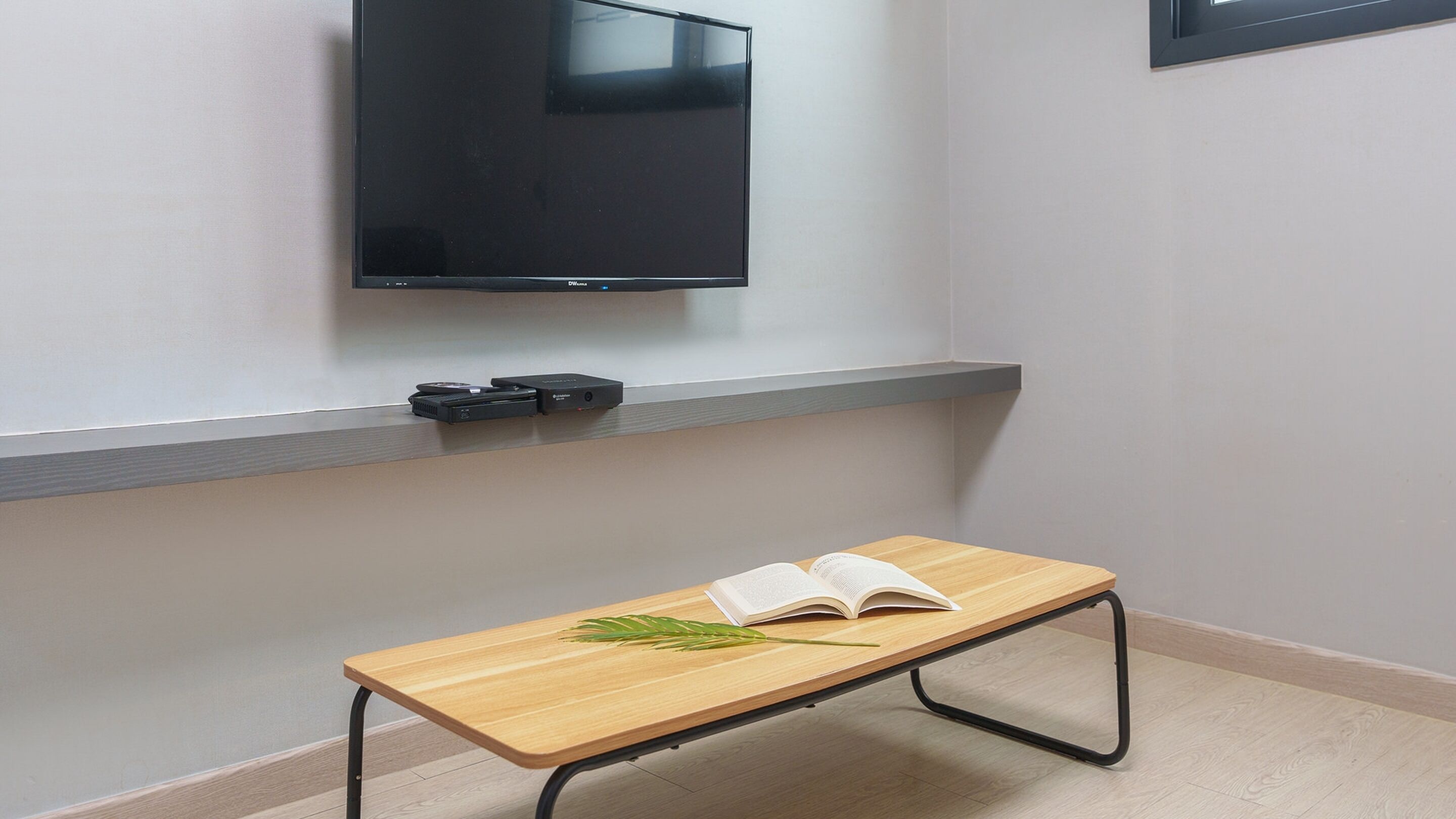 A small wooden table with an open book and a green plant on it, positioned in front of a wall-mounted TV.