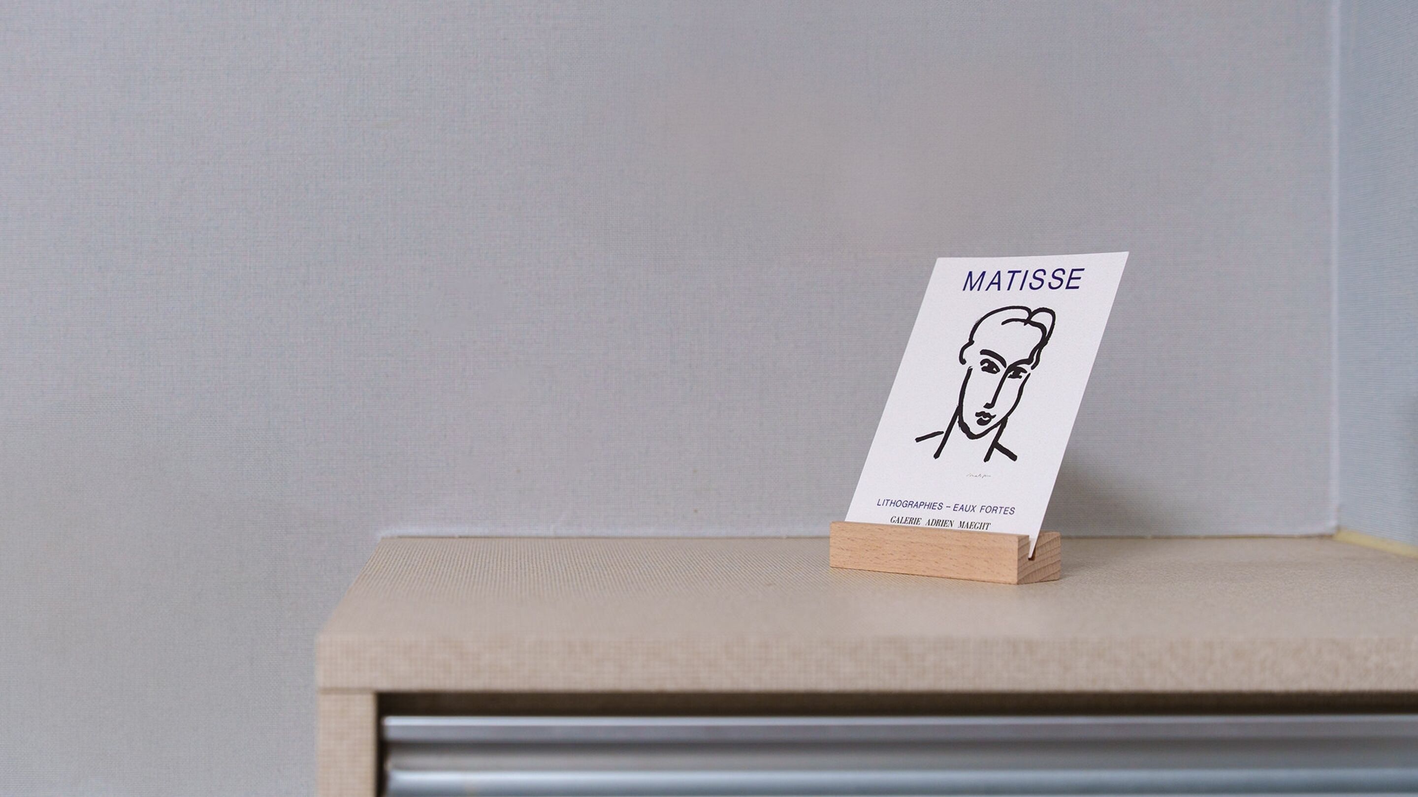 A minimalist display stand with a card featuring a line drawing and text, placed on a shelf against a plain wall.