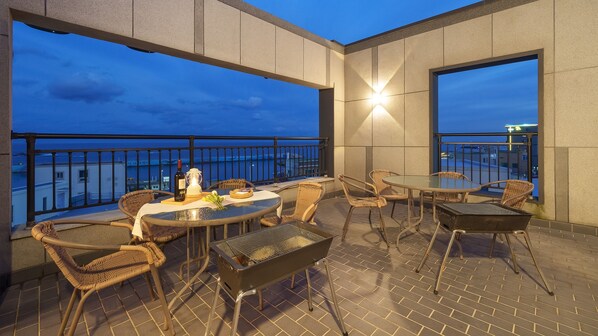 A rooftop terrace with wicker furniture, a grill, and a bottle of wine, overlooking a cityscape at dusk.