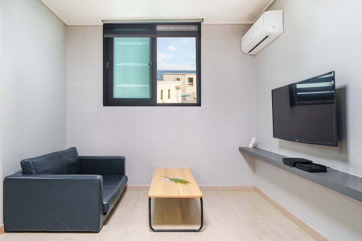 Een moderne, compacte leefruimte met een bank, een klein tafeltje, een flatscreen televisie en een raam met uitzicht op gebouwen.