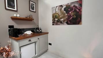 Une cuisine équipée d’une machine à café, d’un évier et d’un plan de travail en bois. Deux tableaux encadrés sont accrochés au mur, au-dessus du plan de travail.