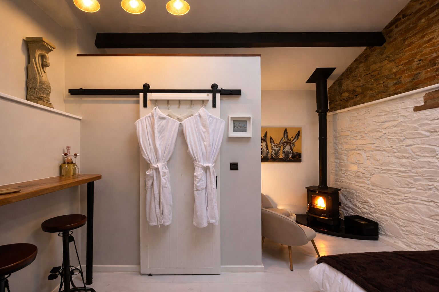 A modern bathroom with a white bathtub, a wooden vanity, a towel rack with two white bathrobes, a stone wall, and a wood-burning stove.