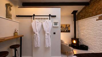 A modern bathroom with a white bathtub, a wooden vanity, a towel rack with two white bathrobes, a stone wall, and a wood-burning stove.