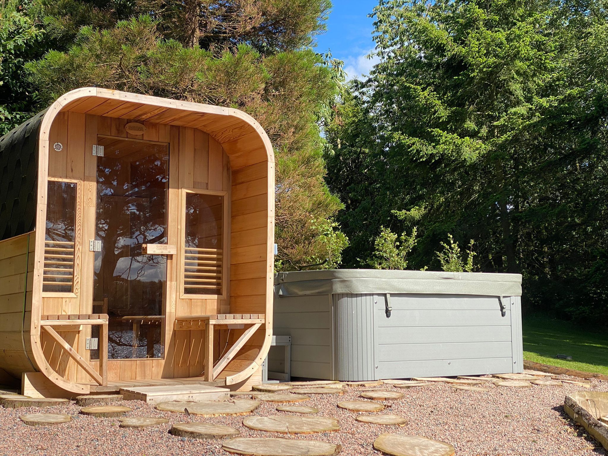 Una cabaña de madera con un ventanal grande y un porche pequeño, rodeada de árboles y un camino de grava.