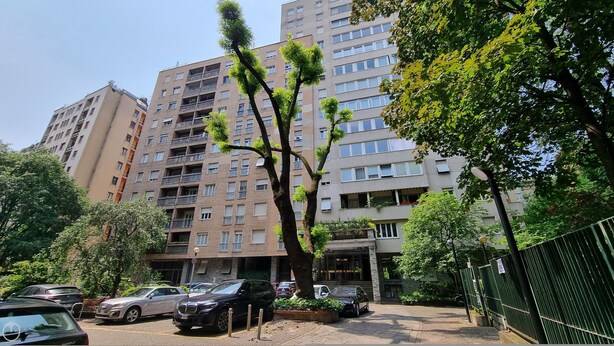 Un edificio residencial de varios pisos con balcones, un estacionamiento con automóviles y zonas verdes circundantes.