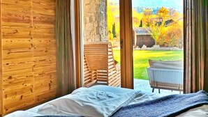 A bedroom with a wooden wall, a bed with a blue blanket, and a view of a stone wall and garden through a glass door. - Alpine Mountain Chalet (Marebbe)