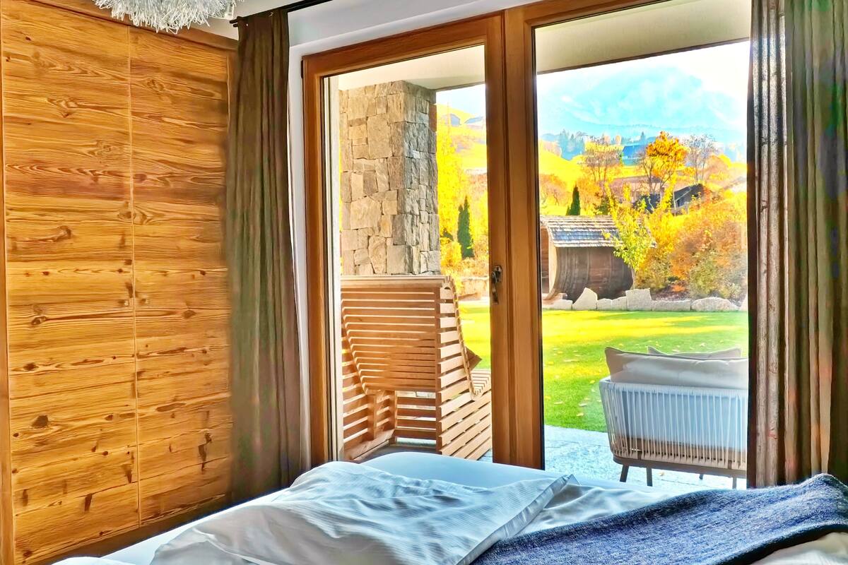 A bedroom with a wooden wall, a bed with a blue blanket, and a view of a stone wall and garden through a glass door.
