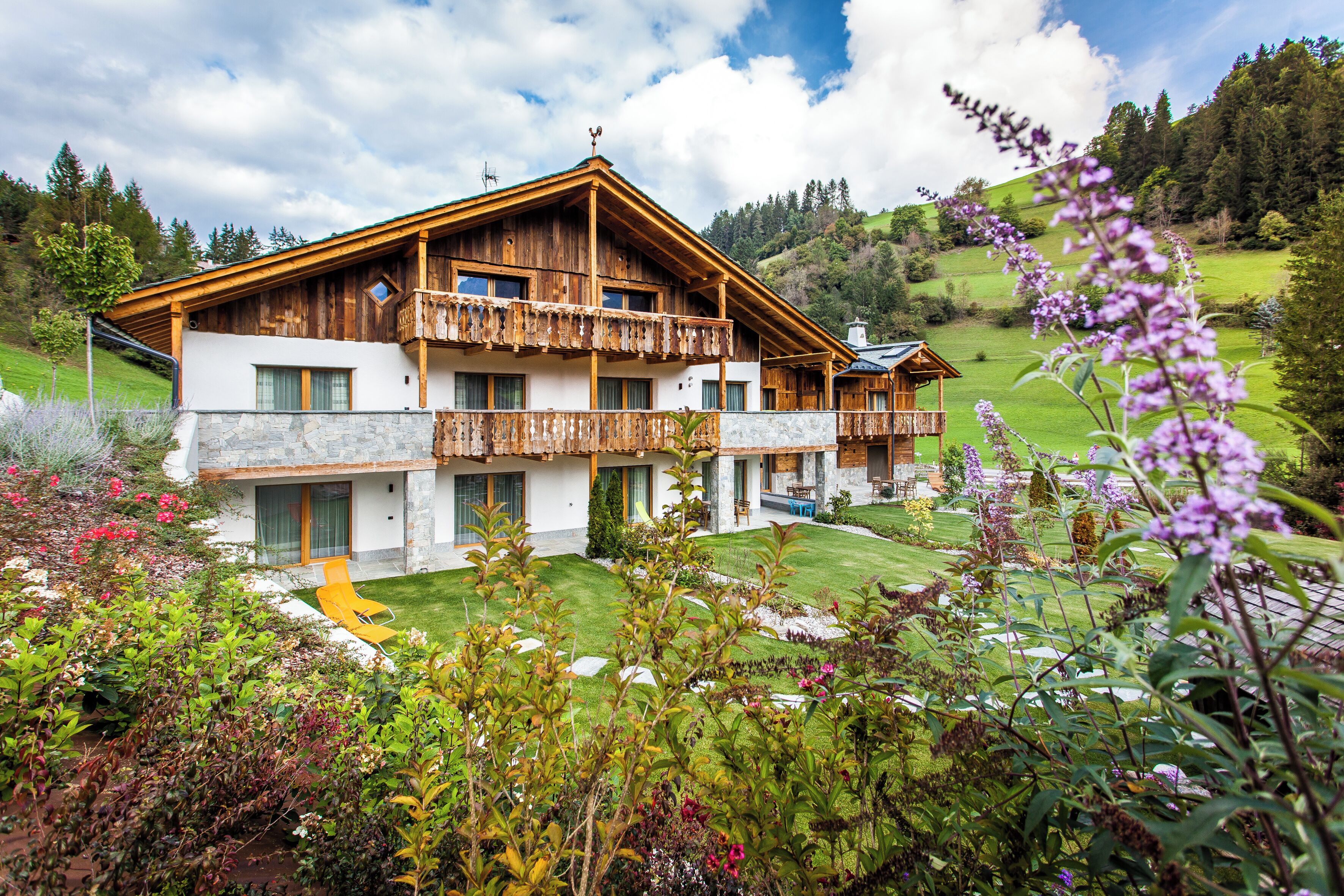 A two-story wooden chalet with a balcony, surrounded by lush gardens and a hilly landscape.