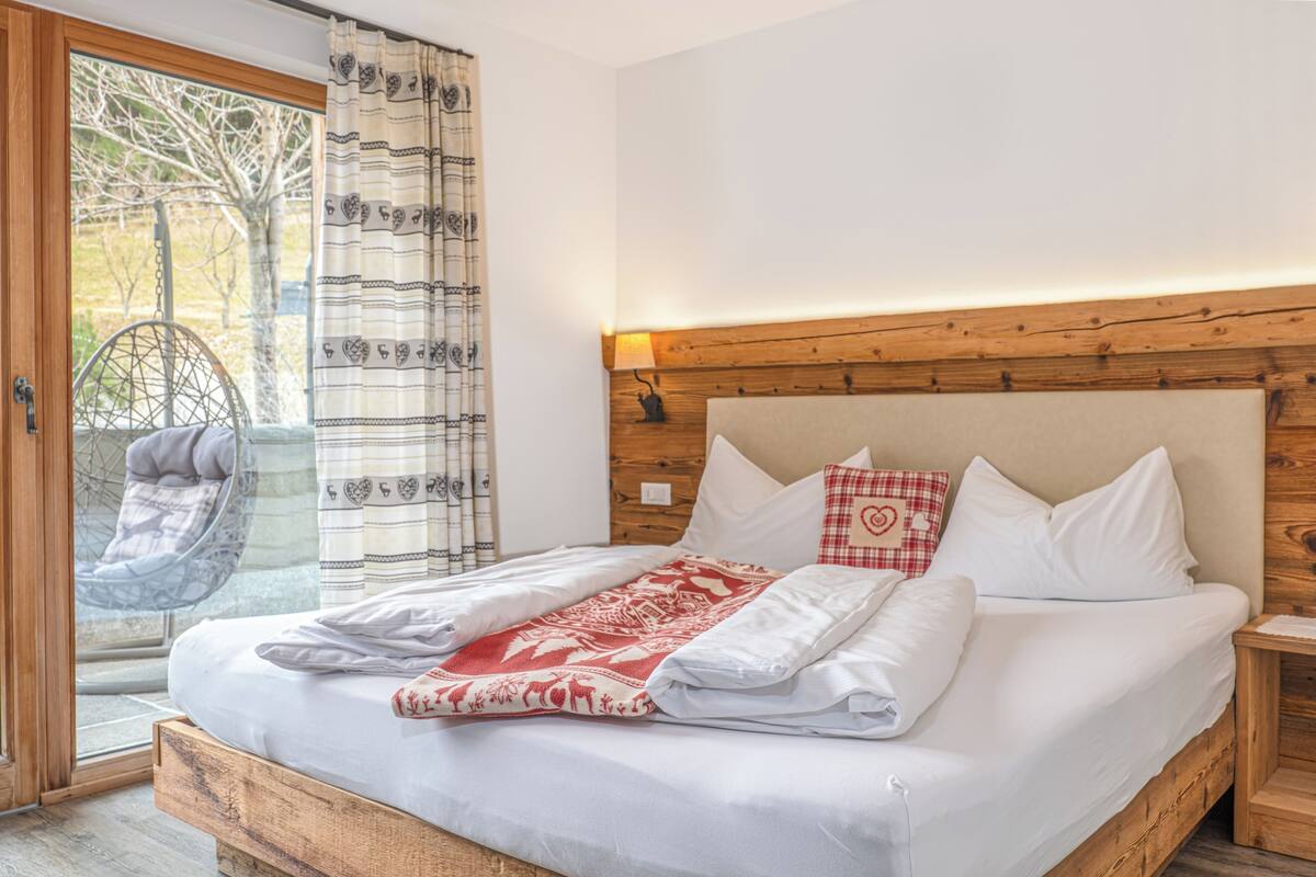 A bedroom with a wooden headboard, a bed with white bedding, a red and white patterned throw blanket, and a chair by the window.